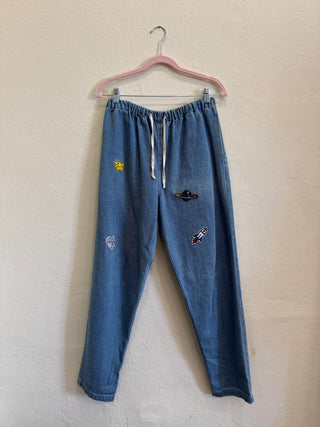 A pair of blue denim draw-string pants with embroidered space-themed patches (a yellow sun, a black UFO, and a rocket) hanging from a pink hanger against a textured off-white wall.
