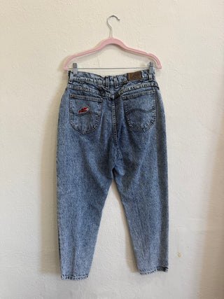 Light blue washed denim jeans shown from the back, hanging on a pink plastic hanger against a textured off-white wall.