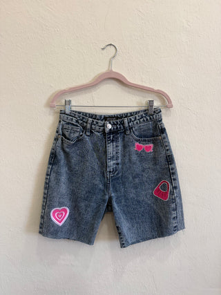 A pair of blue denim shorts with pink embroidered patches (a heart, sunglasses, and a handbag) hanging from a pink hanger against a textured off-white wall.