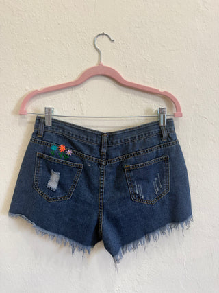 Rustic blue denim shorts with a raw frayed hem, distressed details, and colorful embroidered flowers on the back pocket, hanging on a pink hanger against a textured off-white wall.