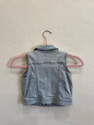 A light blue denim vest hanging on a pink hanger against a textured off-white wall.