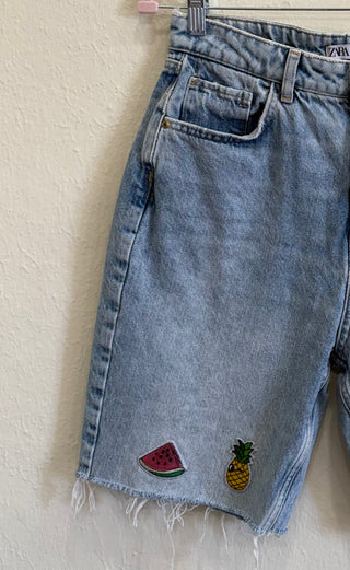 Light wash denim shorts with a raw frayed hem, featuring embroidered patches of a watermelon slice and a pineapple on the lower left leg.
